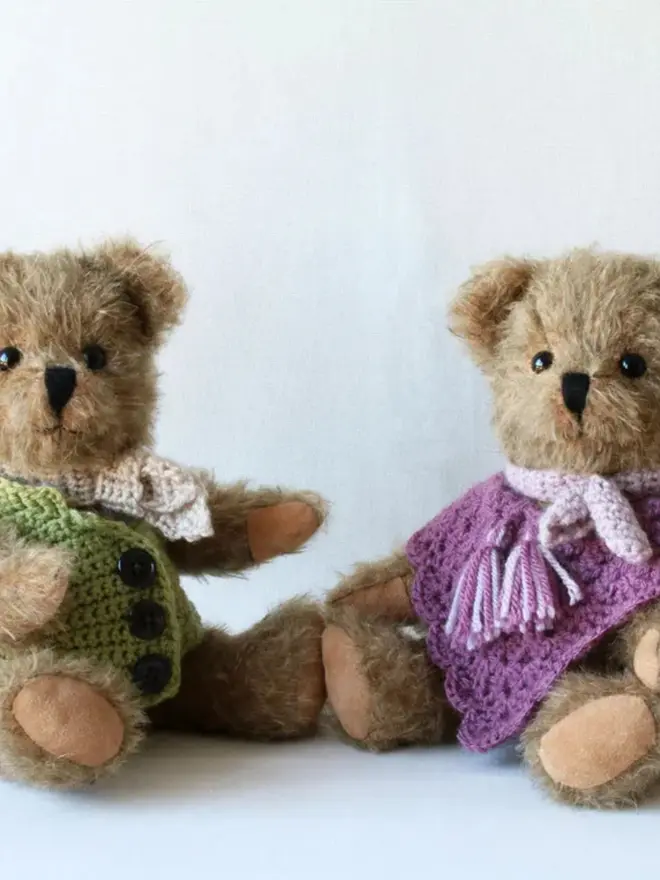 Handmade brown teddy bear wearing a green cardigan and white scarf, sitting with other teddy bears against a plain background.