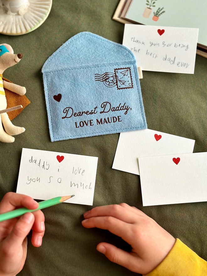 A handmade blue felt envelope with the personalisation 'Dearest Daddy, love Maude' and a paper aeroplane stamp with postmark, lays on dark green fabric. A young child is writing notes to Daddy on the included heart notecards.