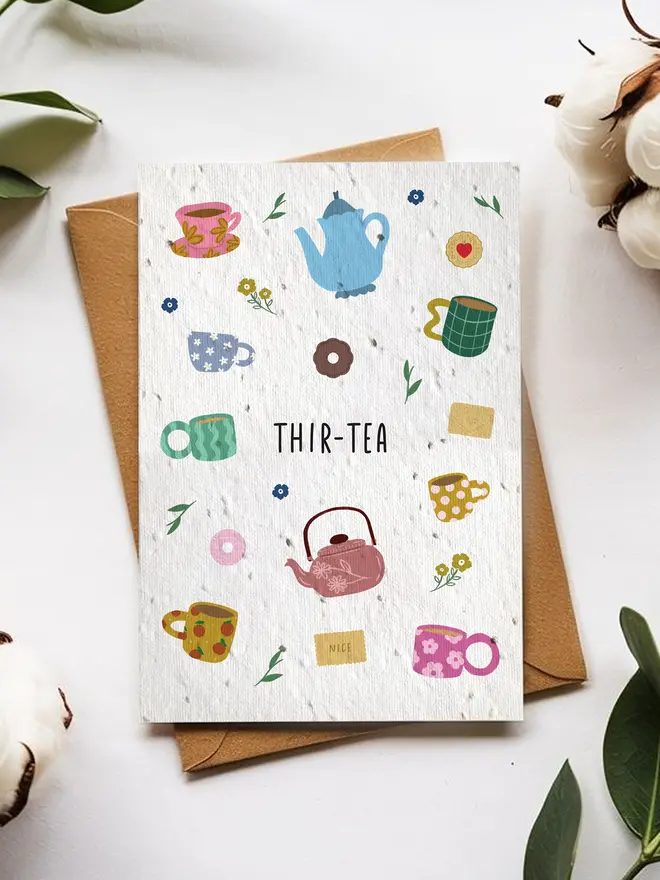 A greeting card titled “THIR-TEA” featuring colorful illustrations of teapots, mugs, flowers, and tea bags, placed on top of a brown envelope surrounded by cotton flowers and green leaves on a white background.