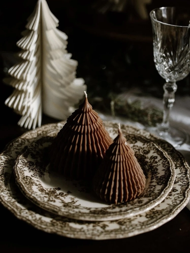 Brown Origami Christmas Tree Warm Candles (Set Of 2). Two brown cone tree candles in different sizes on a floral transfer-print plate, beside a white paper honeycomb tree and a cut-crystal glass. The rich chocolate tone brings a cosy, vintage-inspired feel to holiday dinners.