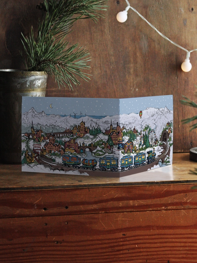 fold out greeting card with christmas town illustration on wooden box with foliage behind