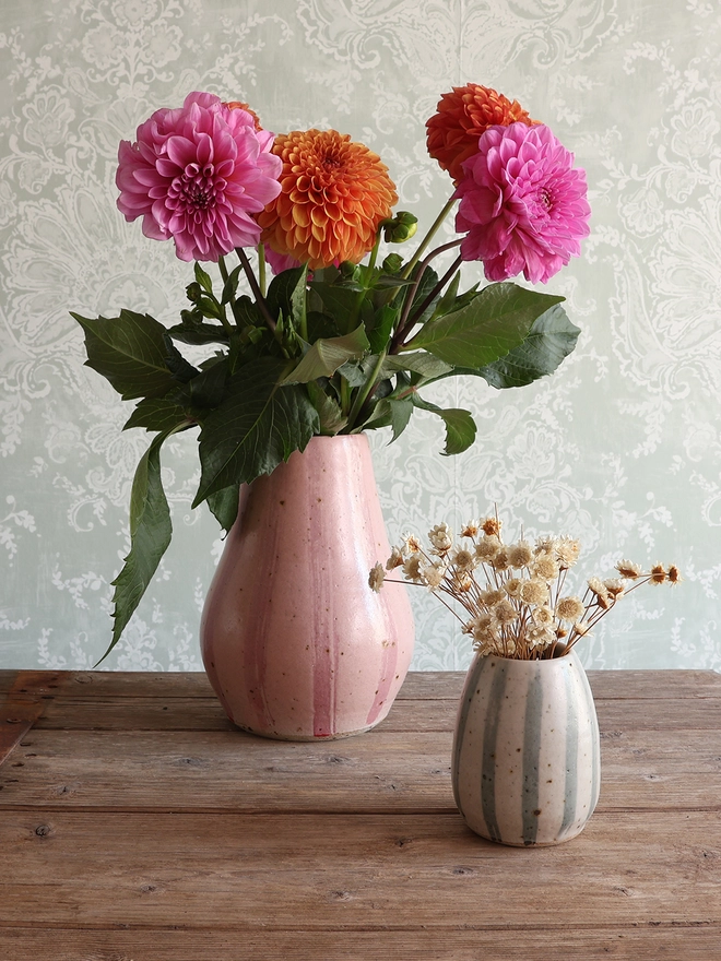 Handmade Ceramic Stoneware Large Vase in Pink Stripe filled with a bouquet of orange and pink flowers sat on a wooden surface. A Small Handmade Ceramic Stoneware Vase with a blue and white stripe design and filled with dried flowers sits in front of it. 