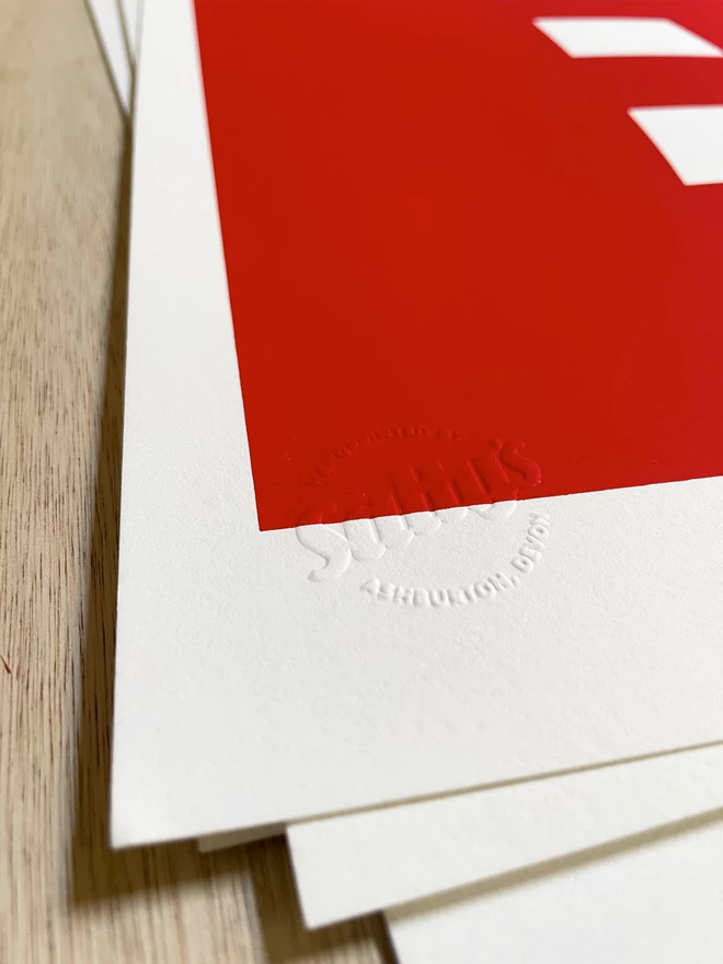 Bottom left corner of a screenprint, showing the Salty’s embossed stamp is in focus in the corner and the red image is receding towards the back of the frame. Laid on a wooden table top.