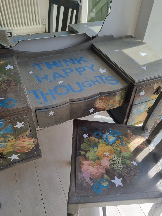 Vintage painted dressing table and chair. Painted in coffee grey and decorated using layered antique florals with yellow and blue accents and finished with hand-cut white paper stars. Text reads in blue 'think happy thoughts'. Heavily distressed finish.