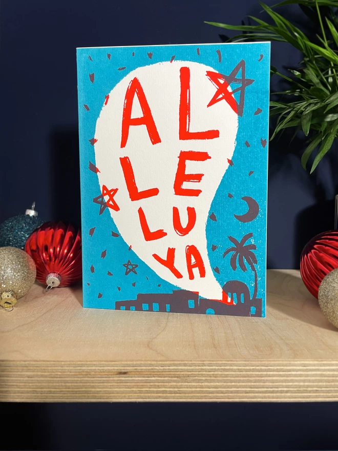 Al-le-lu-ya handprinted Christmas card in turquoise and red, sat on a plywood shelf with baubles and greenery about.
