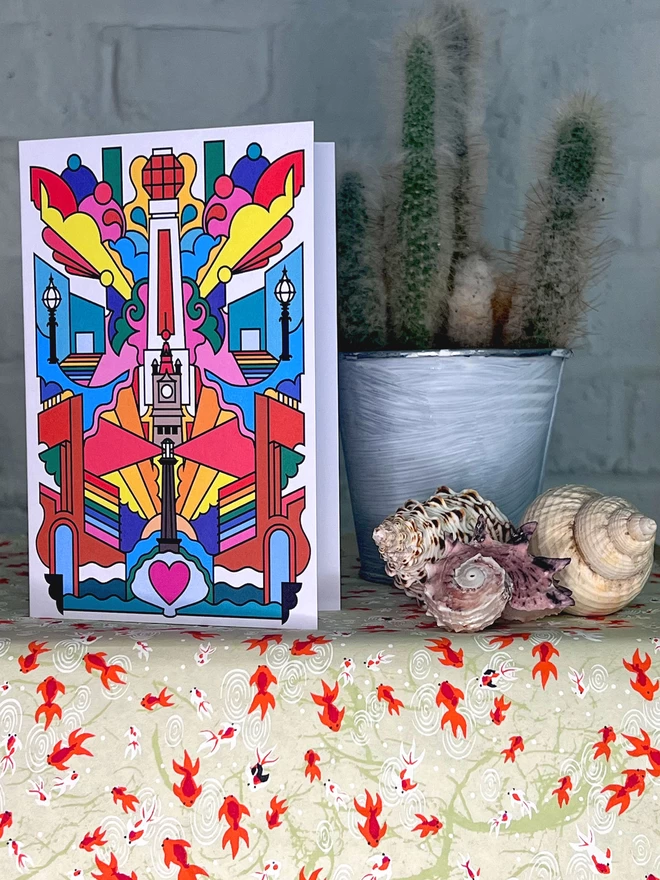 A greetings card of black line drawing with rainbow colours depicting Margate landmarks, sits on a shelf next to a cactus in a silver pot behind sea shells. 
