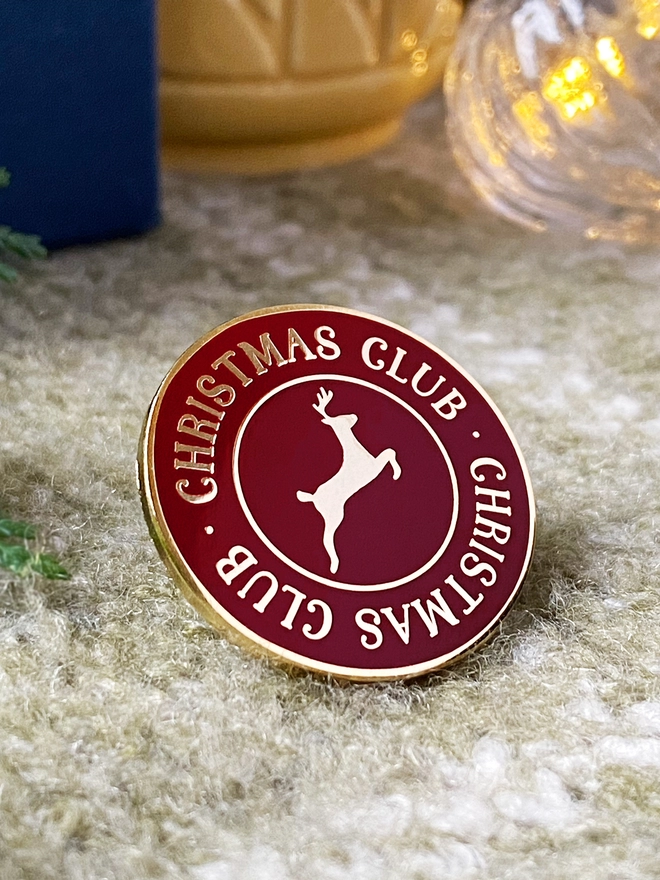 A deep red and gold enamel pin badge with a gold reindeer in the centre and the words "Christmas Club" around the outside is on a green blanket.