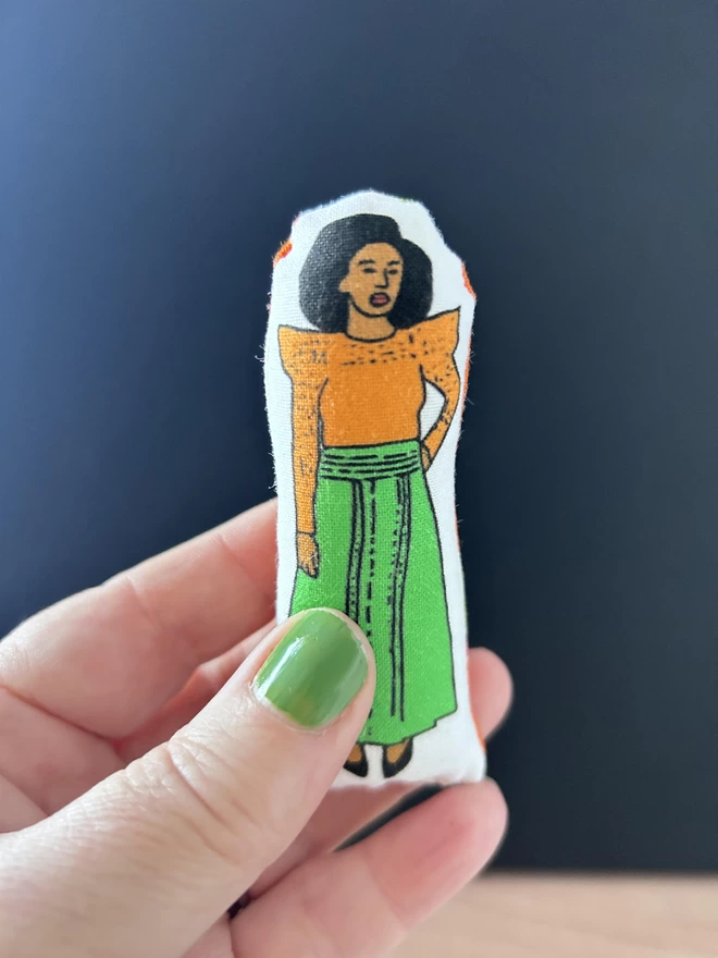 Mini fabric doll of Chimamanda Adichie, wearing an orange top and green skirt held up in a hand against a dark backdrop.