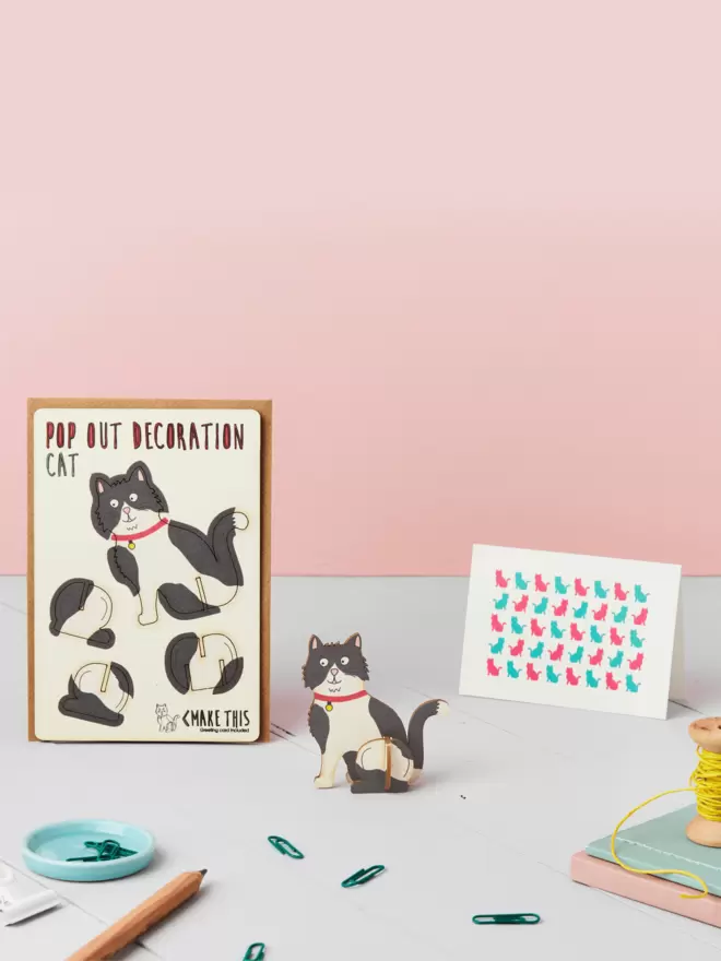 3D black and white cat decoration and cat pattern greeting card and brown kraft envelope on top of a grey desk in front of a light pink coloured background