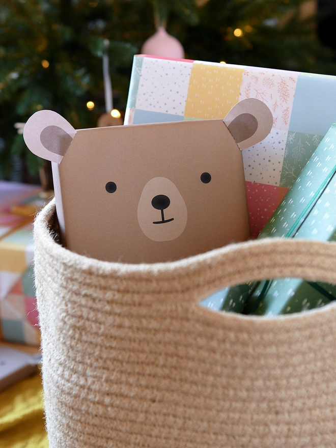 A gift wrapped up as a brown bear is tucked into a woven basket beside other wrapped gifts in front of a Christmas Tree.