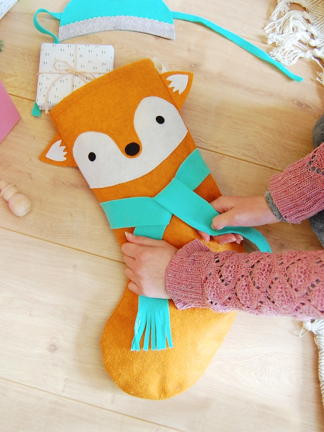 A handmade orange felt fox Christmas stocking lays on a wooden floor while a green scarf is being tied around it.