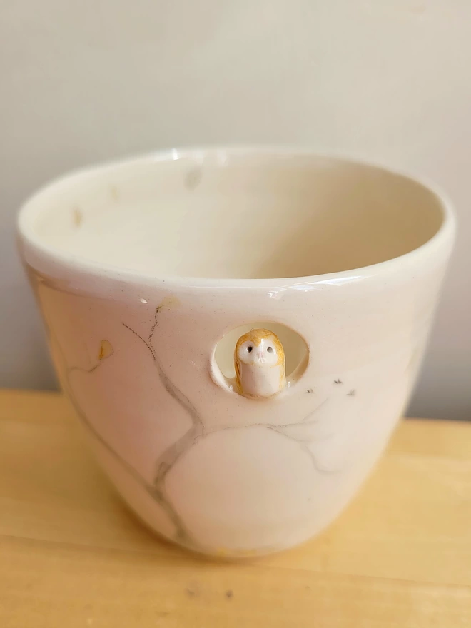 cream ceramic cup on a wooden surface with an owl perching in a cut out at the top of the drinking vessel with hand painted tree, leaves and bird prints