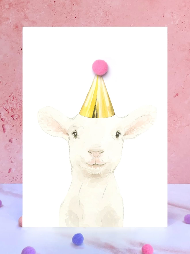 A greeting card featuring a hand painted design of a lamb, stood upright on a marble surface surrounded by pompoms. 