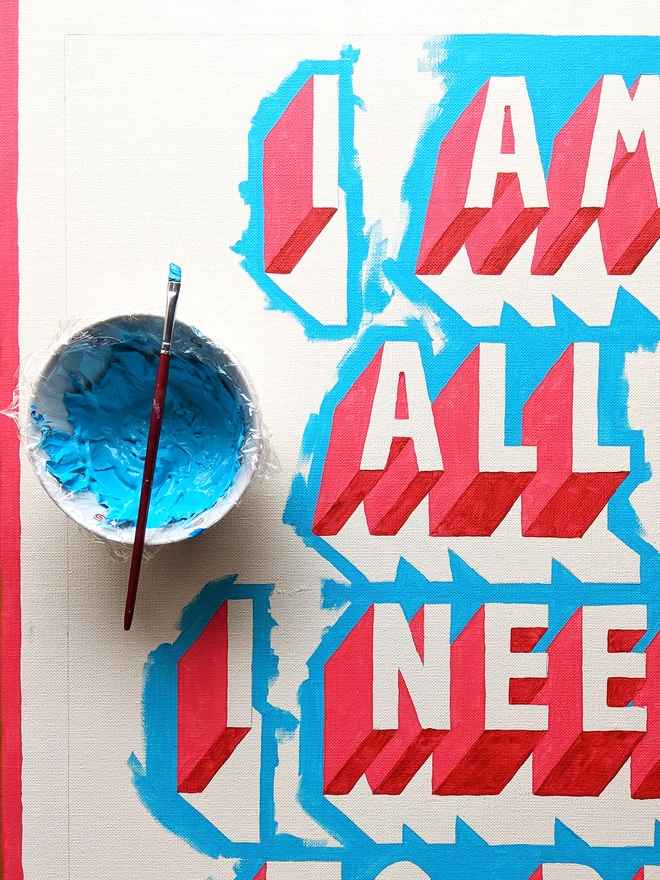Overhead view of a partially painted typographic artwork by artist Survival Techniques reading “I AM ALL I NEED TO BE.” A blue paintbrush rests on a pot of blue paint beside the artwork. Some letters are fully painted while others are still in progress, revealing the layered process behind the bold 3D design in red, white, and blue on a blue background.