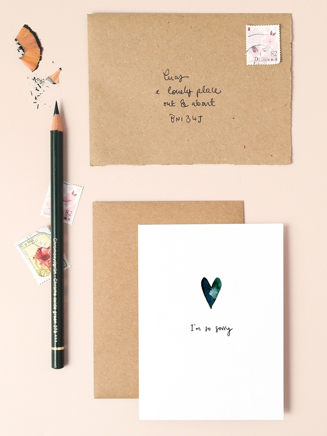 An image of a mini, rectangular white greetings card with an illustration of a dark blue heart with a hand written 'I'm so sorry' message in black ink seen under the central illustration. The card is seen on top of a brown rectangular kraft envelope.