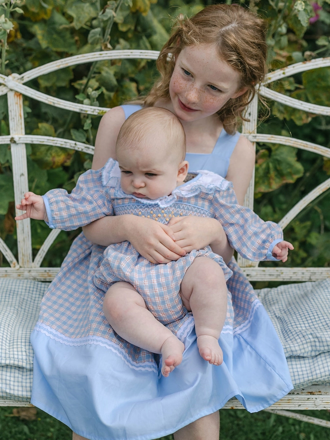 A girl holds a baby while sitting on a bench. They are both wearing matching checked clothes. the girl in a dress and the baby in a romper.