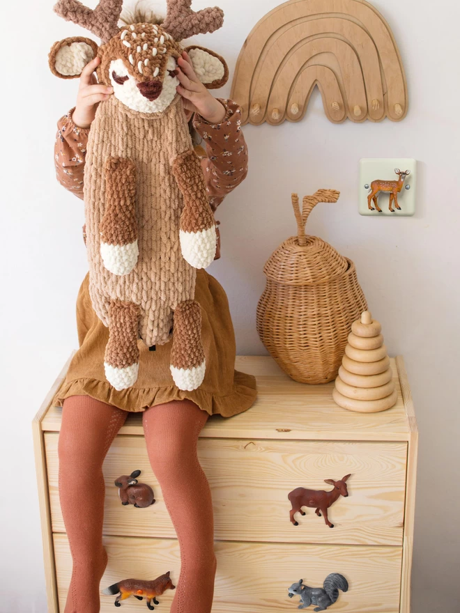 A pine chest of drawers with four woodland animal drawer knobs attached, the animals include a brown rabbit, a brown deer, a fox and a grey squirrel. A 5 year old girl sits on the drawers, she is dressed in brown vintage clothes and holds a woodland fawn toy over her face. On the wall their is a brown wooden rainbow, a woodland fawn light switch by Candy Queen Designs and a wicker basket in the shape of a pear. 