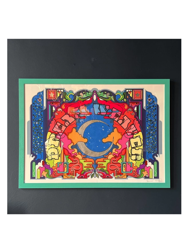Framed in a teal, hung on a dark grey wall, the image a silver waxing crescent moon sits in the centre under the words “Dream Weaver” which arches around it. The central illustration is brightly coloured, and is edged with a dark blue border.