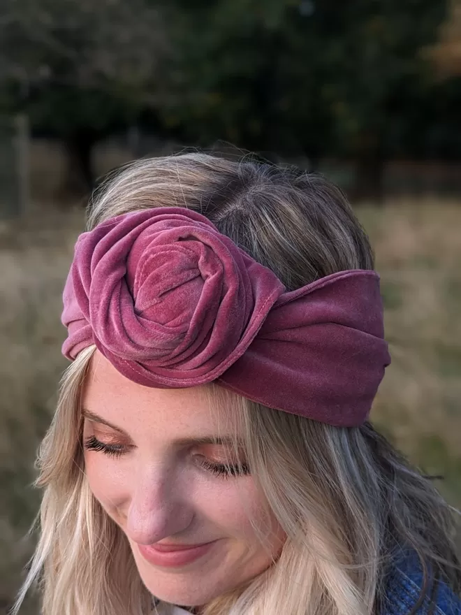A dusty rose colour headband made from velvet fabric, tied in a rose-like knot on a blonde-haired woman.