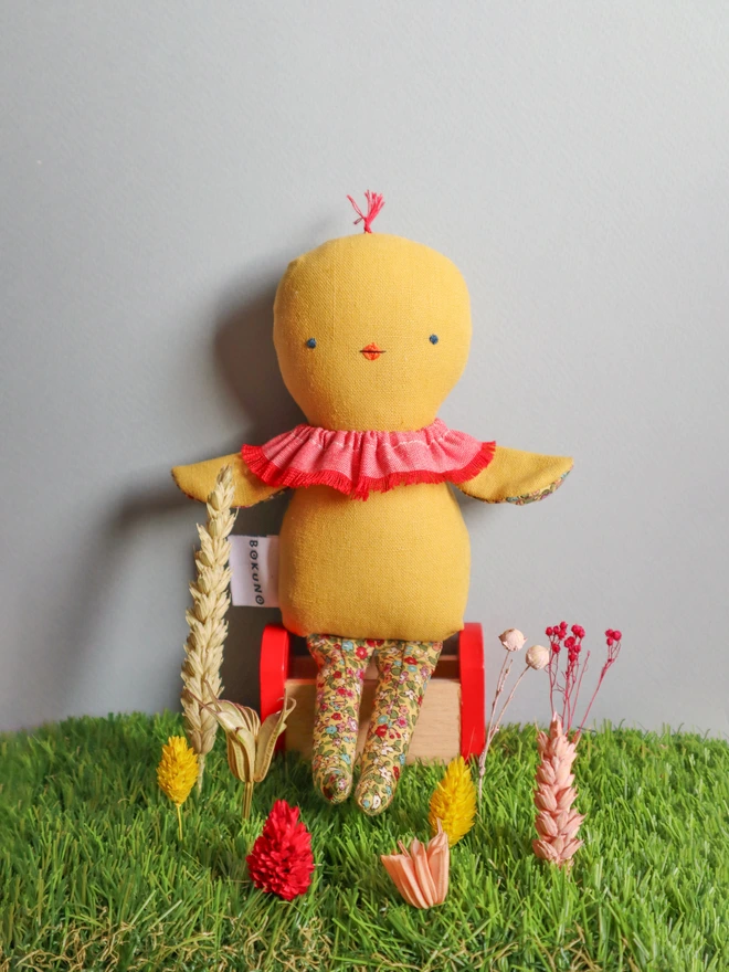 Handmade chick doll made from soft yellow fabric, with a stitched face, small beak, ruffled collar, and patterned fabric arms and long legs, sat on a grass with flowers.