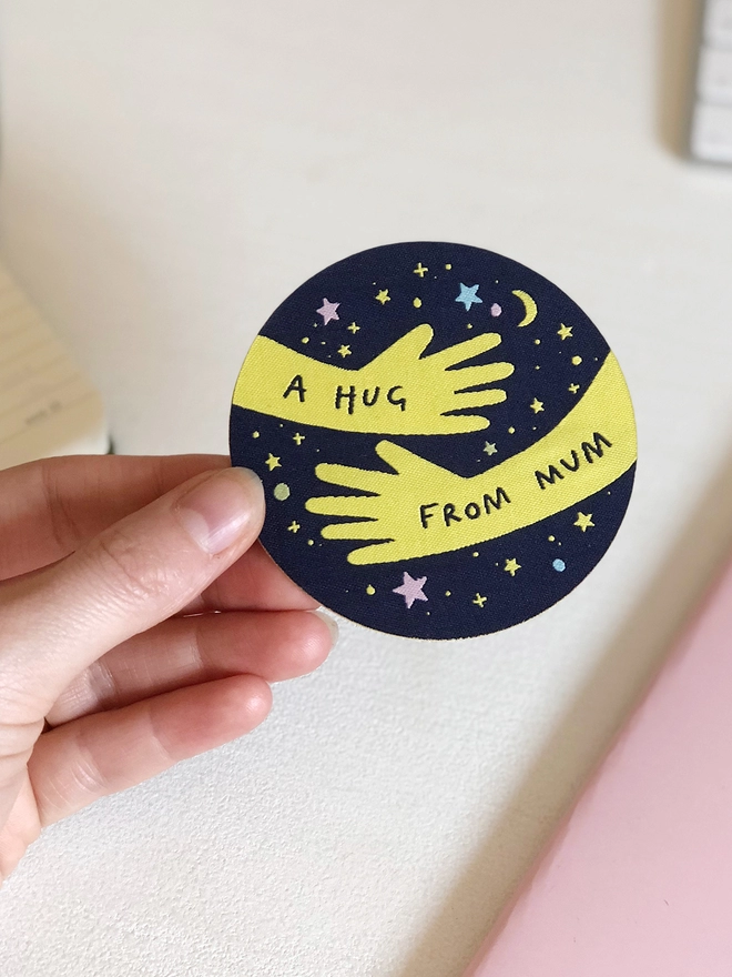 A navy blue and yellow woven patch with two arms in a hugging position and the words "A hug from Mum" is being held in a hand.