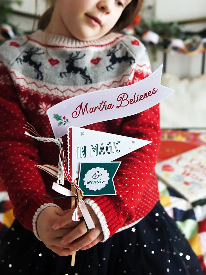 A child wearing a traditional red Christmas jumper holds three personalised Christmas flags made from cardboard. Each flag is attached to a wooden stick with ribbons. The flags read "Martha Believes in magic & wonder".