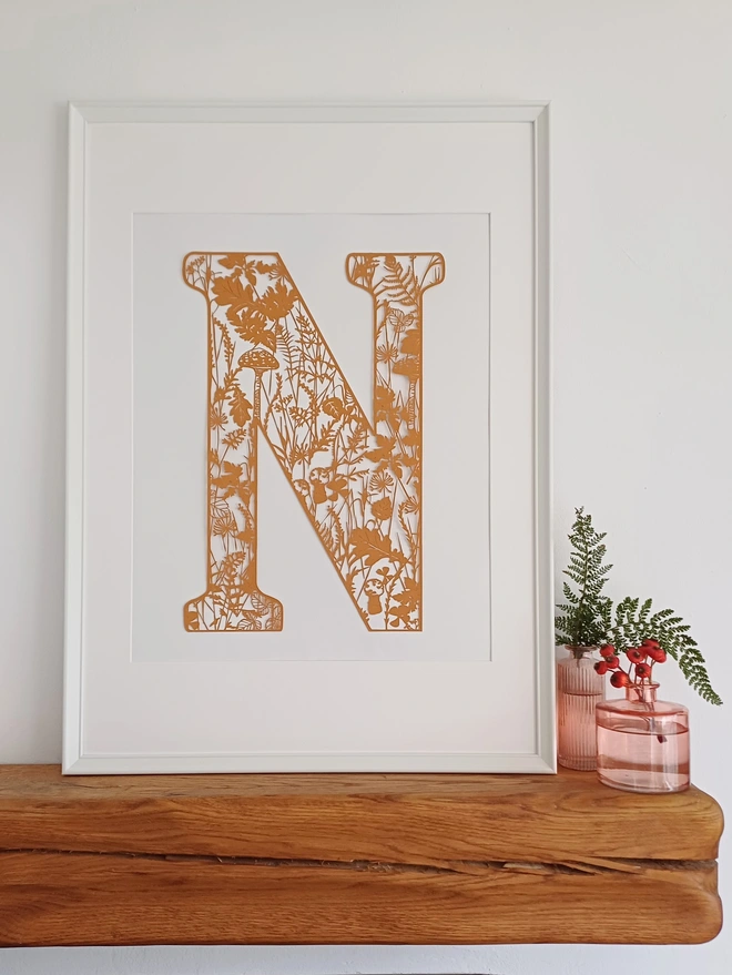Framed Letter N papercut in raw sienna paper with detail of autumn plants, flowers and fungi