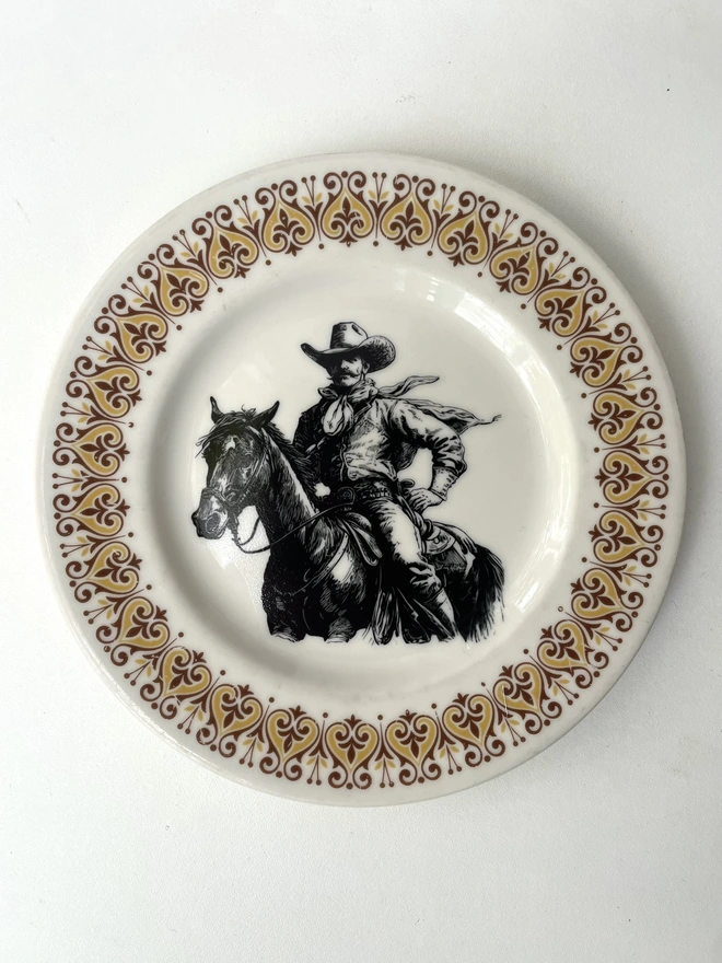 vintage plate hanging on a white wall, it has a decorative border, with a printed vintage illustration of a cowboy in the middle 