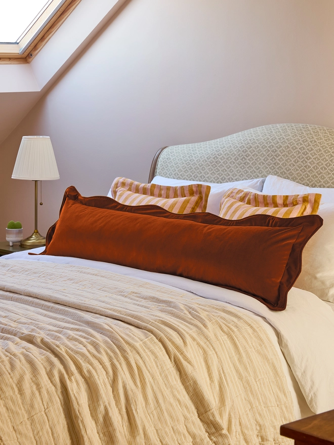 Handmade scalloped rust velvet bolster cushion 130cm with feather pad paired with stripe linen cushions on bed, Kika Bespoke