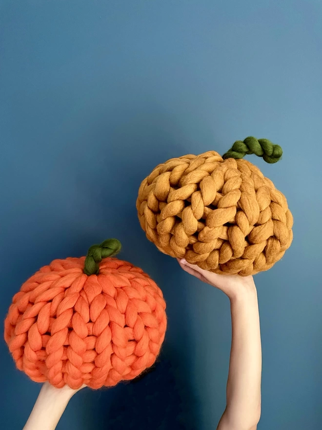 Two knitted pumpkins in orange and yellow seen held up in front of a blue wall.