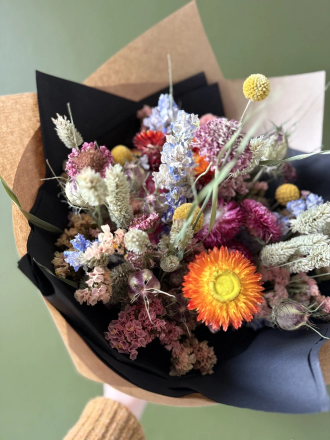 a dried flower bouquet in bright colours wrapped in black tissue and kraft paper held by a person in a mustard coloured jumper with their left arm against a green wall