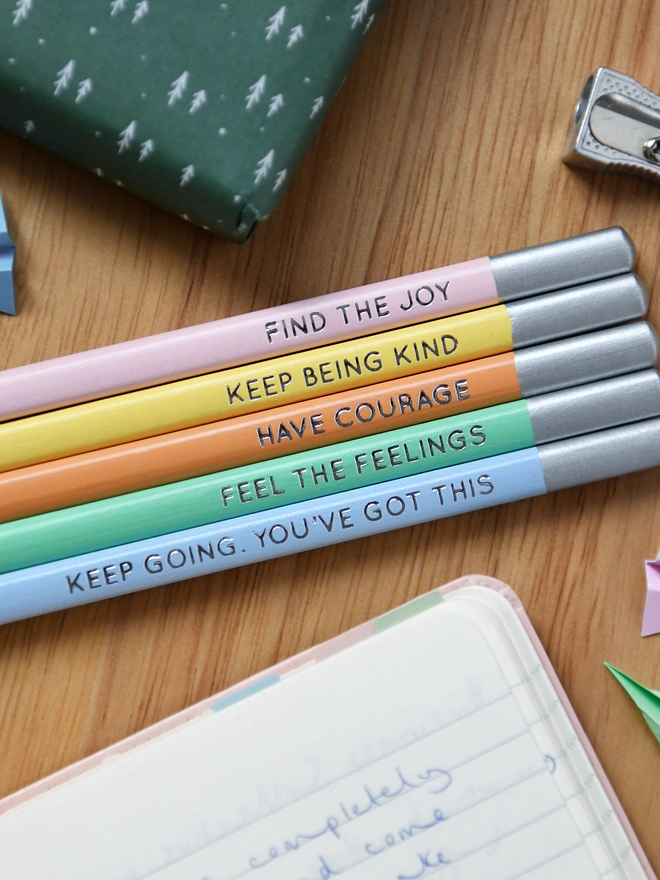 Five pastel coloured pencils placed on a wooden desk next to a silver pencil sharpener. Each pencil has a positive message stamped in silver block foiling.