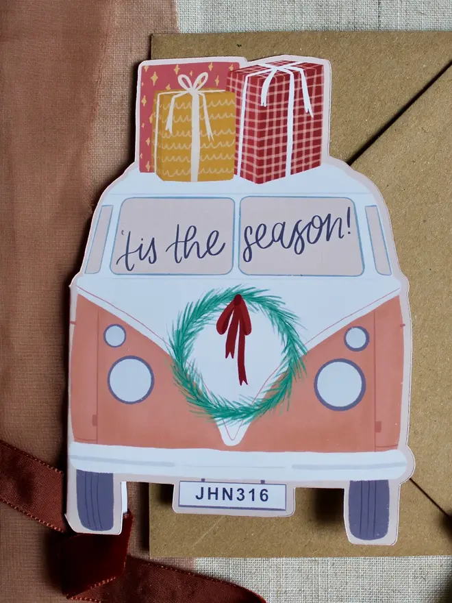 tis the season illustrated campervan christmas card. pink illustrated campervan card emblazoned with phrase tis the season and decorated with christmas gifts and a wreath.