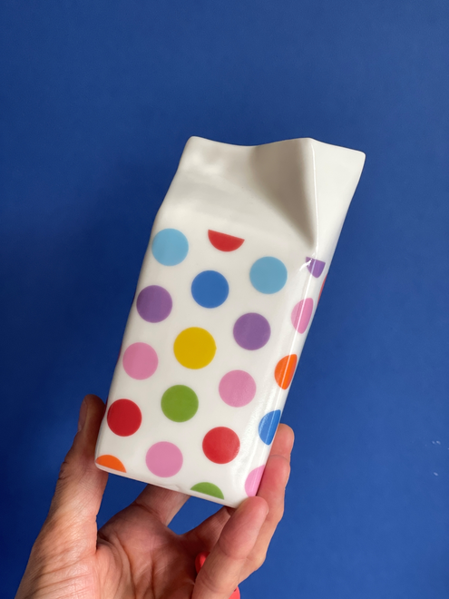 Hand holding milk carton jug on a blue background , the jug has a multicoloured polkadot pattern in red , blue , green , orange , purple and yellow 