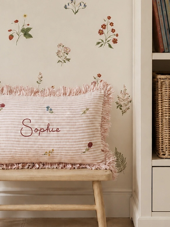 Personalised pink striped stonewashed linen cushion with ruffle trim, hand-painted ditsy florals and embroidered name, styled in a floral children’s bedroom.