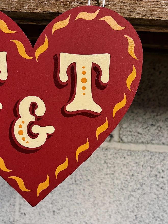 Close-up detail of a wooden love heart cut out with a red matte background, glossy golden yellow curve details painted along the edges and ivory lettering with dark red shade.