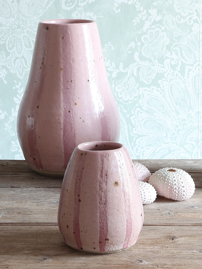 Handmade Ceramic Stoneware Small Vase in Pink on a wooden table. Behind the vase sits a large vase in the same style and a collection of three pink sea urchins. 