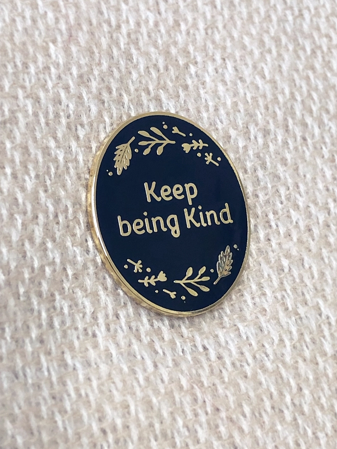 A navy blue and gold enamel pin  with the words Keep Being Kind with a floral border is pinned onto a white scarf.