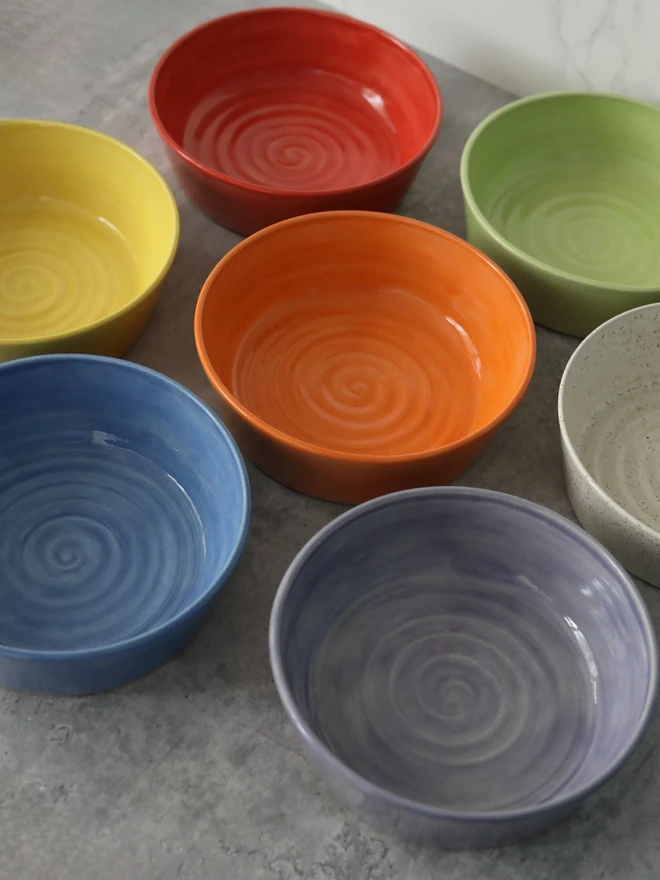 Seven colourful glazed ceramic bowls arranged on a grey surface, viewed from above at an angle, in yellow, red, orange, green, blue, lavender, and speckled white, each featuring a swirl pattern on the interior.