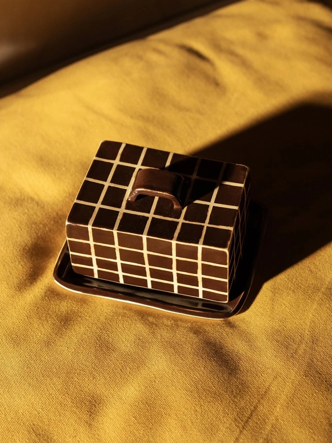 Grid Butter Dish, a colourful butter dish with a grid pattern sitting on a yellow bed surface. 