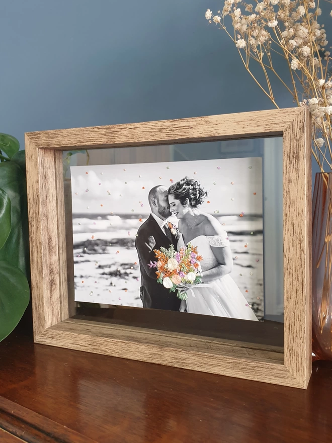 Wedding photo with hand embroidered bouquet and confetti held in double glass frame on desk