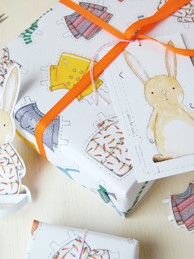 A gift wrapped in white wrapping paper with a hand illustrated outfit design and rabbit paper doll gift tags is beside a cut out rabbit paper doll on a white wooden table.