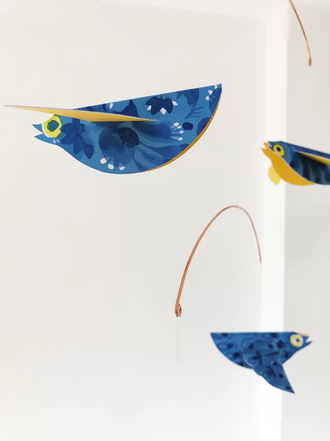 Three bright blue patterned paper birds mobile hanging against a white wall. They show sunshine-yellow bellies.