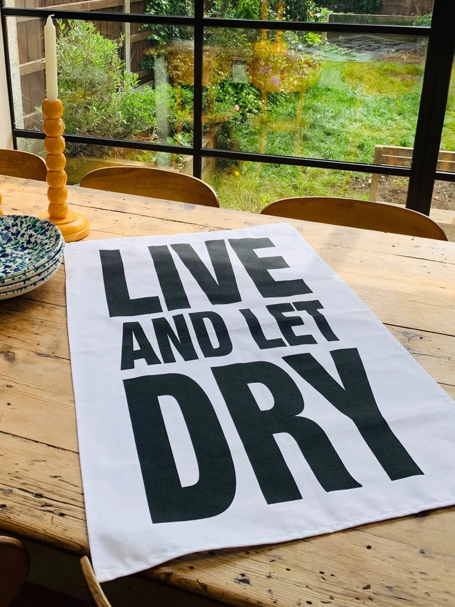 London Drying Live and Let Dry black screen printed text on white tea towel laying on timber table with stack of dishes and candlestick on table. Garden seen through glass panes in background