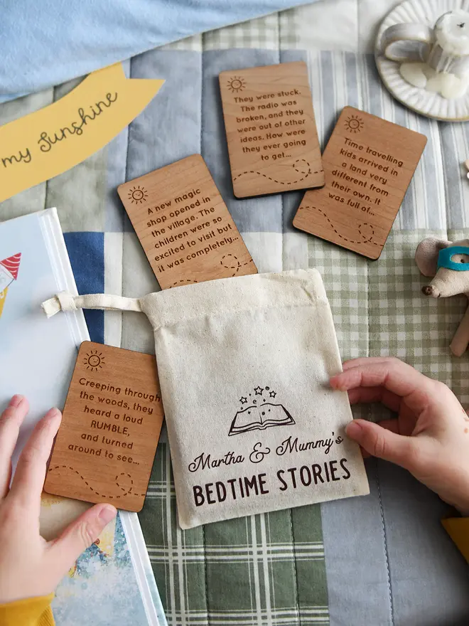 Personalised bedtime story set for dads and children featuring engraved wooden story prompt cards and a drawstring cotton bag reading 'Martha & Mummy’s Bedtime Stories', displayed on a patchwork quilt with a soft toy, picture book, and a young child holding one of the cards.