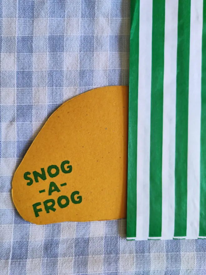 back of hand painted cornish pasty drawing, with green and white striped paper bag, sitting on gingham cloth