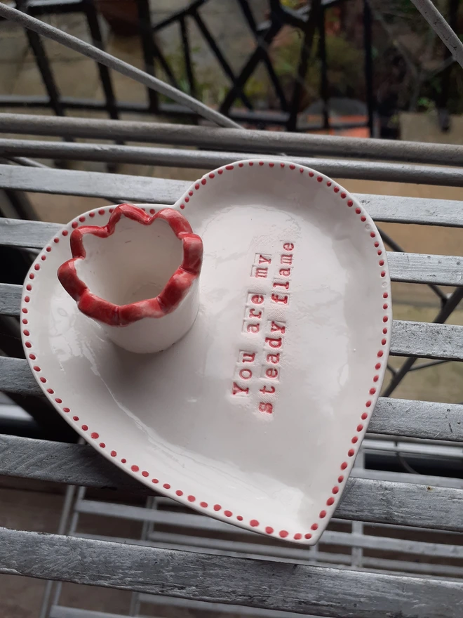 Romantic handmade ceramic candle holder shaped like a heart, with red trim and stamped lettering reading ‘You are my steady flame’, photographed on a rustic metal table.