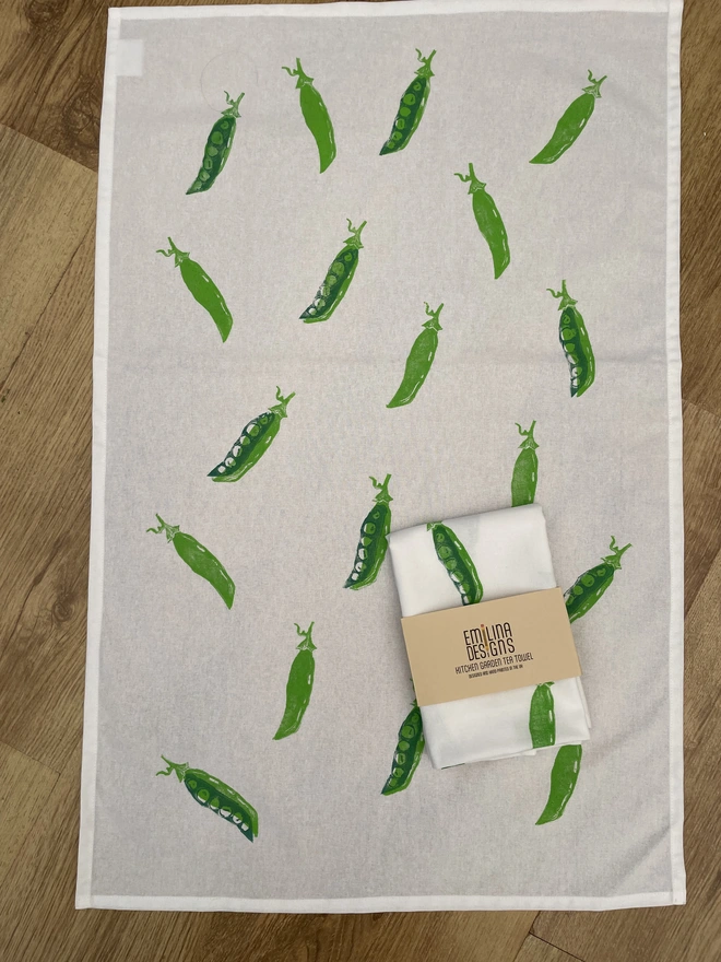 Pea Kitchen Tea Towel. White fabric with a diagonal pattern of green pea pods, some open to show bright green peas.