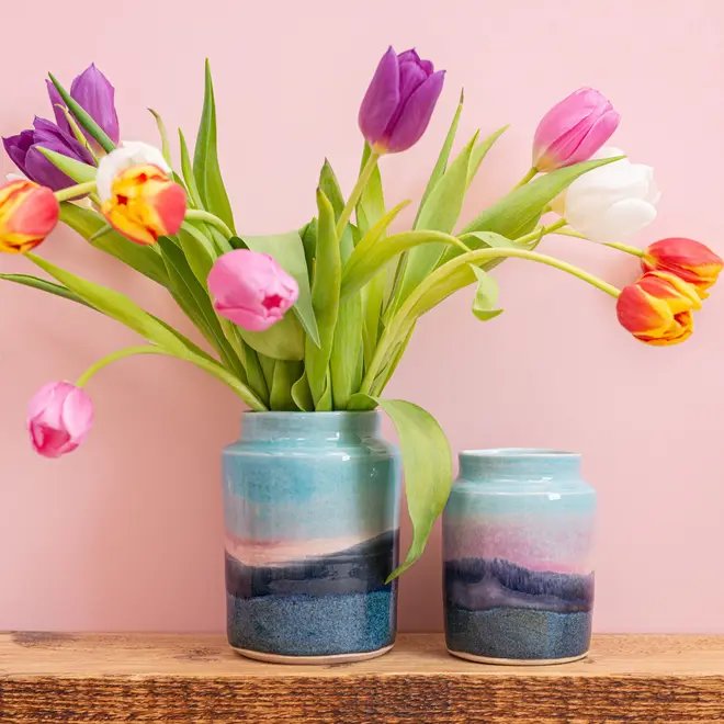 Landscape Medium Vase, a pair of colourful vases sitting on a wooden surface against a patterned backdrop. They are accompanied by some flowers. 