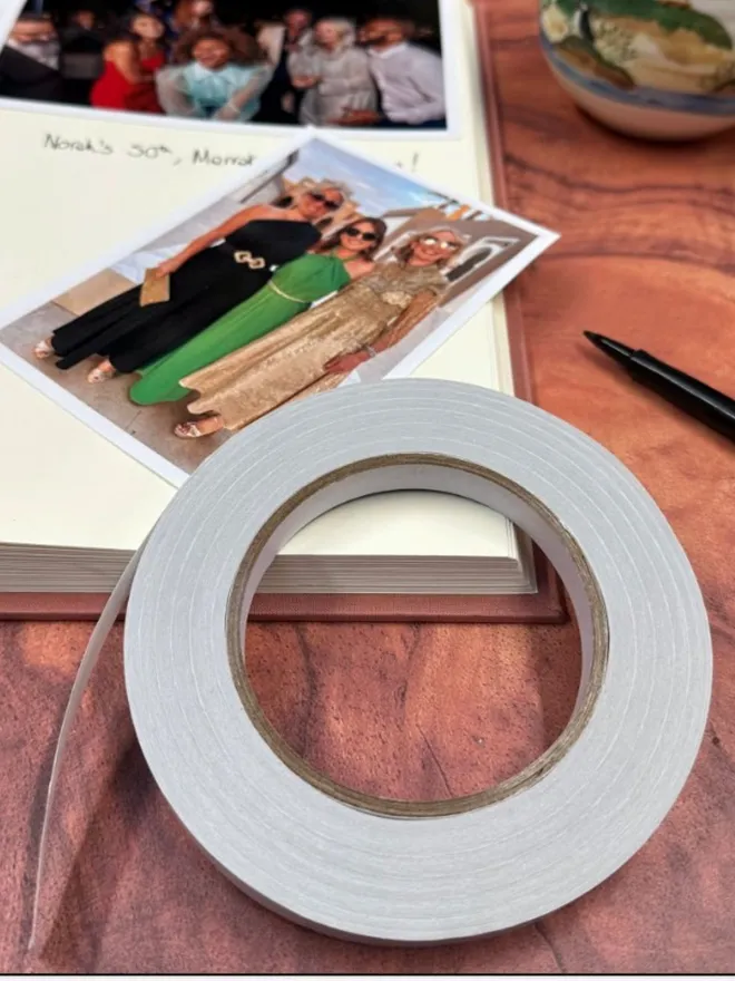A roll of photo mounting tape is resting on a wooden surface and the page of an open photo album.